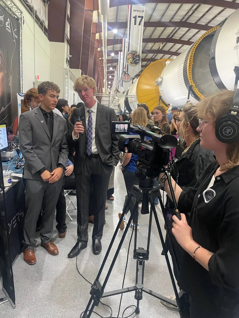 Sam Iannone being interviewed on camera at the NASA HUNCH event at Johnson Space Center, with a Saturn V rocket and Apollo 17 banner behind him