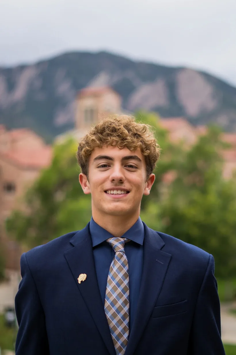 Headshot of Sam Iannone in a navy suit on the CU Boulder campus with the Flatirons behind him